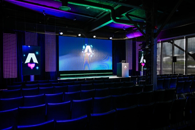 View from the back of the dark empty keynote presentation area. Rows empty black chairs. A vibrant large LED screen with the Astro Together logo on it. Two smaller portrait-oriented screens adorn the sides of the stage, also with the Astro Together logo.