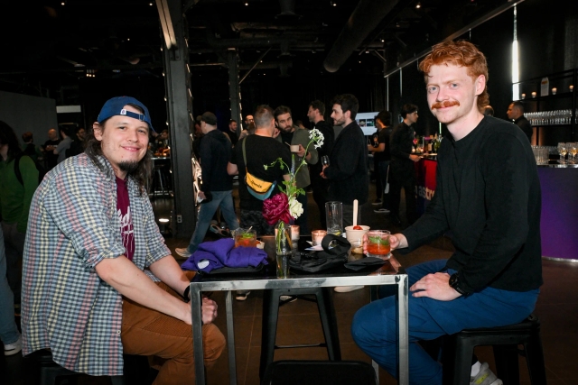 Two male guests pose sitting at a high table with their food, drinks, and apparel.