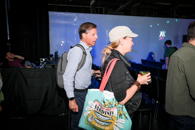 Two older guests walk into the presentation area with their bags and drinks.