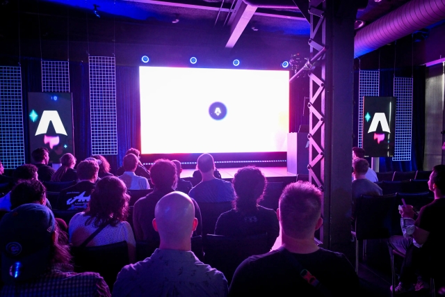 View from the back of guests in their seats, waiting for the keynote presentation to begin. They are brightly lit in purple by the large LED screen on stage.