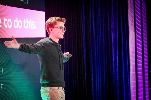 A close up of Ben Holmes on stage with both arms outstretched, gazing at the audience. He is lit with pink and purple lighting.