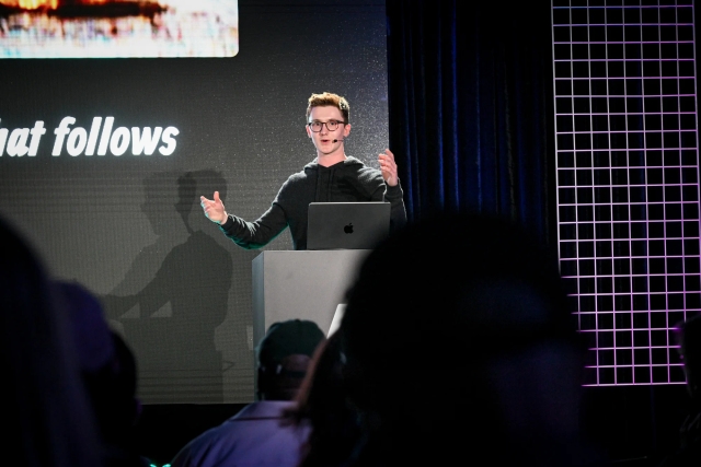 Ben Holmes speaking from an Astro-branded podium. Both his hands are up as he addresses the audience.