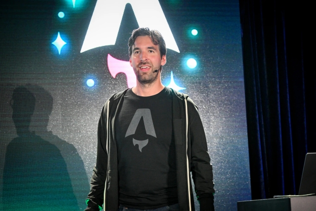 Fred K. Schott speaking on stage, gazing at the audience. Behind him is a large Astro Together logo.