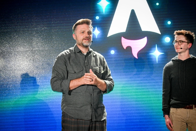 Closeup of Matthew Phillips standing with other keynote speakers on stage as he answers an audience question. Off to the right, Ben can be seen looking at Matthew.