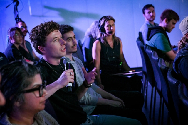 An audience member holding a microphone, asking keynote speakers a question. A member of the A/V team can be seen watching the audience member intently.